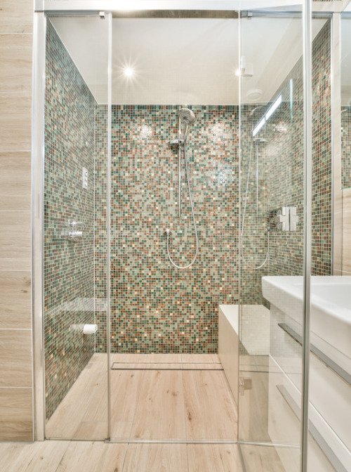 The image displays a bathroom shower in Fort Mill featuring multi-colored mixed squares glass pool tiles. The tiles are arranged in a repeating pattern, creating a visually stunning and unique look. The mix of colors and shapes adds texture and visual interest to the space, while the glass material creates a sense of transparency and reflection. The overall design is modern and sleek, with clean lines and minimalist fixtures that create a sense of openness.
