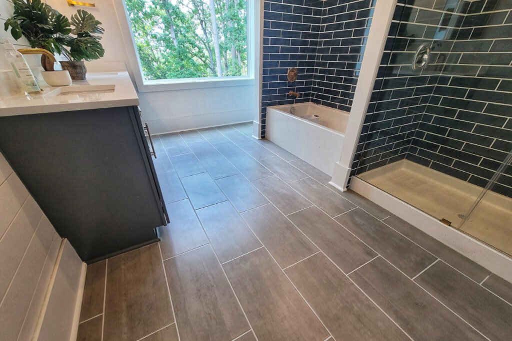 The image shows a recently remodeled bathroom in Fort Mill, SC. The flooring is composed of grey tiles that are laid out in an offset pattern. The shower and tub area is tiled with blue subway tiles, which give the space a modern and stylish look. The overall design is clean and contemporary, with sleek fixtures and a neutral color palette.