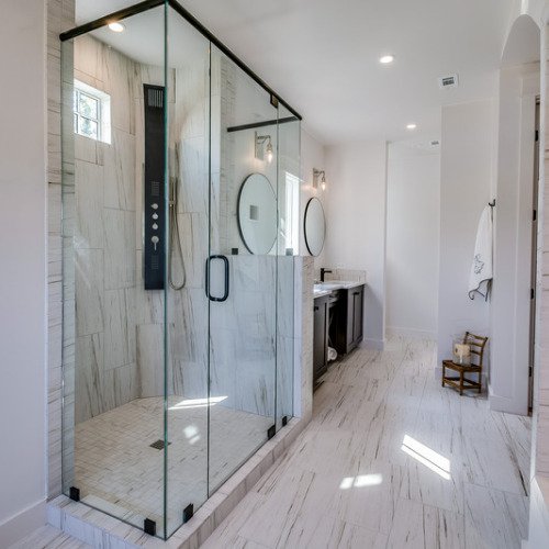 Image of a modern shower in Fort Mill, SC, featuring an all-glass enclosure and a sleek shower system. The shower is located in the foreground and the walls are covered with tiles that match the bathroom floor, featuring a mix of tan, gray, and white shades. The contemporary design of the shower provides a clean and sophisticated look to the bathroom.