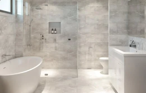 Image of a beautifully remodeled warm bathroom in Fort Mill, SC. The bathroom features uniform warm light gray floor and wall tiles that provide a sleek and modern look. The bathroom includes a free-standing tub located next to a glass-enclosed shower, adding to the luxurious feel of the space. The elegant design and neutral color palette create a soothing and inviting ambiance, making this bathroom a perfect sanctuary for relaxation.